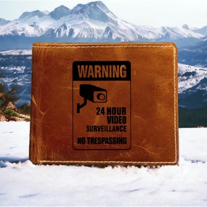 May include: A brown leather wallet with a black warning sign that reads "WARNING 24 HOUR VIDEO SURVEILLANCE NO TRESPASSING". The sign features a black silhouette of a security camera.