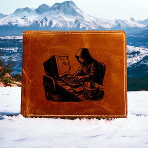 May include: A tan leather bifold wallet featuring a dark brown laser-engraved illustration of a hooded figure sitting at a desk, typing on a vintage computer monitor and keyboard, with a small mug placed next to a stack of books on the desk surface.