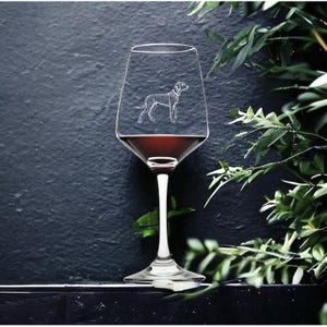 Pode incluir: Uma taça de vinho de vidro transparente com vinho tinto dentro. A taça tem um desenho gravado de um cão no lado.