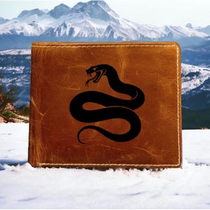 May include: A brown leather wallet featuring a black snake design. The wallet is rectangular with a stitched border. The snake is coiled with its tongue extended. The background shows a snowy landscape and mountains.