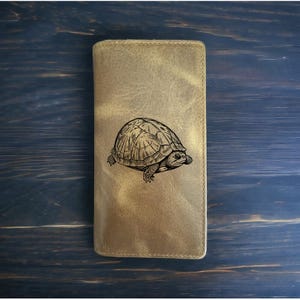 May include: A tan leather wallet with a detailed black illustration of a turtle on the front. The wallet has a rectangular shape and a stitched border. The background is a dark blue wooden surface.