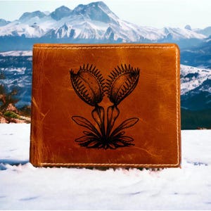 Puede incluir: Una cartera rectangular de cuero marrón con una ilustración grabada en negro de una planta atrapamoscas Venus centrada en la parte frontal. La cartera tiene costuras visibles a lo largo de los bordes y una textura suave y desgastada en el cuero.
