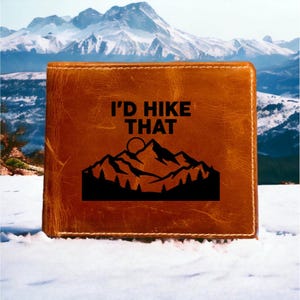 May include: A tan leather bifold wallet featuring a black engraved graphic of a mountain range with a sun and a forest of pine trees. The text I'D HIKE THAT is printed in a bold black sans-serif font above the mountain illustration.