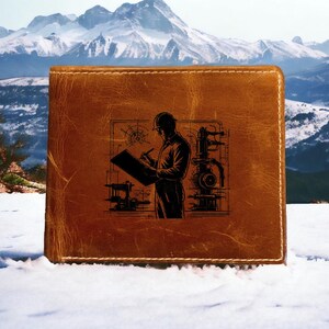 May include: A brown leather wallet featuring a black illustration of an engineer sketching plans. The wallet is rectangular and made of textured leather. The background shows a snowy landscape with mountains.