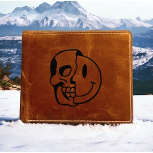 May include: A brown leather wallet with a black skull and smiley face design. The design is half skull and half smiley face.