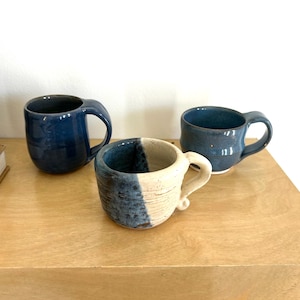 May include: Three ceramic mugs in different styles. One mug is blue with a white stripe, another is blue with a white rim, and the third is a solid blue.