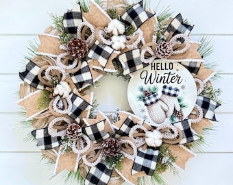 May include: A decorative wreath featuring green pine needles, natural burlap ribbons, and black and white buffalo check ribbons. It is accented with brown pinecones and white cotton bolls. A round sign in the center reads Hello Winter.