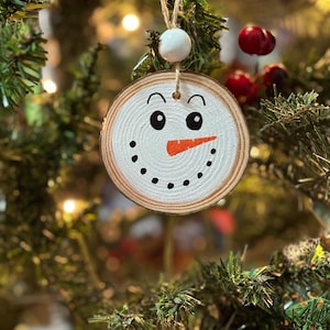 May include: A wooden snowman ornament with a white painted face, black eyes, an orange carrot nose, and black dots for a mouth. The ornament is hanging on a Christmas tree.
