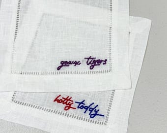 May include: Two square white linen cocktail napkins featuring hemstitched borders. One napkin is embroidered with the purple text geaux tigers, and the other features the red and blue embroidered text hotty toddy in cursive script.