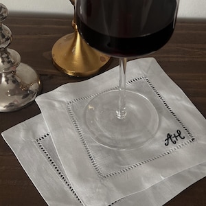 May include: Two white linen coasters with embroidered initials and a decorative stitched border. A wine glass with red wine sits atop one coaster. A silver and gold candle holder are in the background.