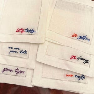 May include: White linen cocktail napkins with embroidered text in various colours. Phrases include "Hotty Toddy", "We are Penn State", "Geaux Tigers", "Go Gators", "Go Dawgs", and "War Eagle". Each napkin has a stitched border.