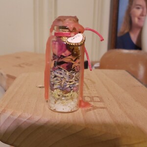 May include: A small glass bottle filled with layers of dried herbs, flowers, and crystals. The bottle is sealed with pink wax and a small heart-shaped charm that says "Love". A pink string is attached to the charm.