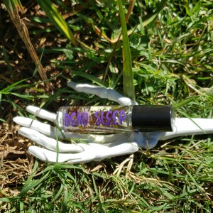 May include: A clear glass roller bottle with a black cap, filled with dried herbs, rests on a white, skeletal hand. The bottle has the words "DEAD SLEEP" in purple lettering. The background is green grass.