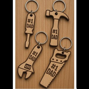 May include: Wooden keychains in the shape of tools: a screwdriver, hammer, wrench, and saw. Each keychain has a silver ring and the text "#1 DAD" engraved on it. The keychains are on a wooden surface.