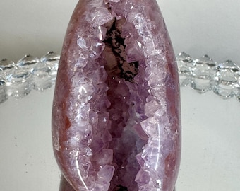 May include: An oval-shaped amethyst geode featuring a deep, hollow center lined with translucent, light purple crystalline structures. The exterior of the stone is smooth and polished with a dark reddish-brown hue, contrasting with the jagged, sparkling interior crystals.