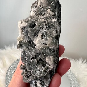 May include: A dark gray crystal cluster with white crystalline formations. The rough, natural stone has a textured surface and is held in a hand. The crystal is a unique, decorative item.