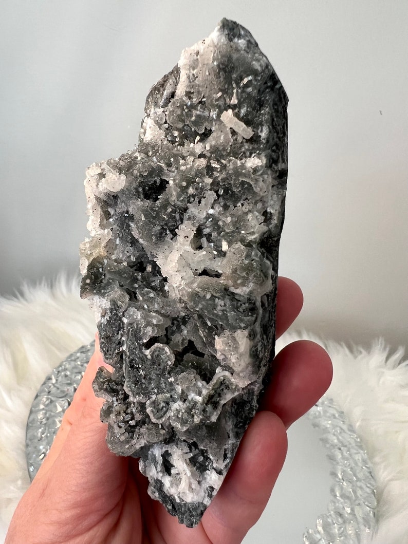 May include: A dark gray crystal cluster with white crystalline formations. The rough, natural stone has a textured surface and is held in a hand. The crystal is a unique, decorative item.