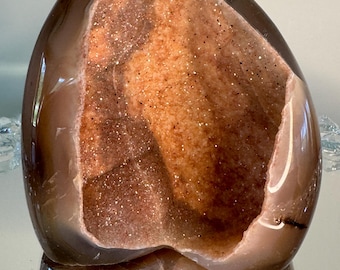 May include: A polished brown agate geode featuring a large, open cavity filled with sparkling, light orange and tan crystalline structures. The smooth, dark brown exterior contrasts with the textured, glittery interior of the mineral specimen.