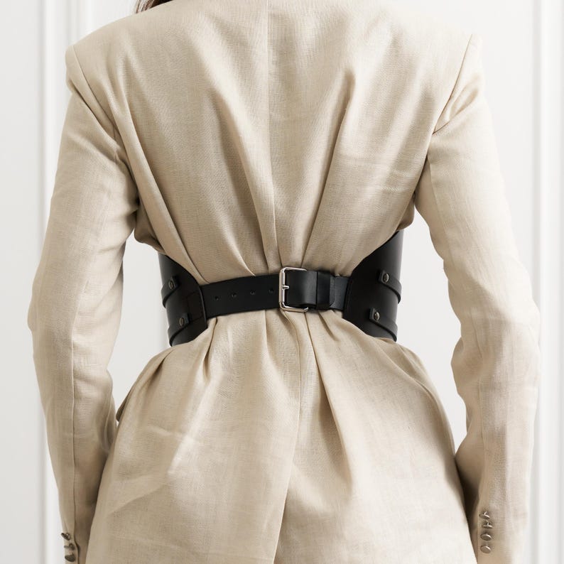 May include: A black leather corset belt with a silver buckle cinches the waist of a beige blazer. The belt has a wide design with multiple straps and rivets, creating a structured silhouette. The blazer has a classic cut with button details.