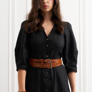 May include: A tan leather belt with two parallel straps and silver buckles. The belt has multiple holes for adjustment and is worn over a black dress. The dress has a V-neck and button-down front.