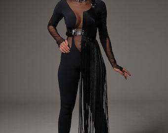 May include: A black jumpsuit with sheer mesh sleeves and bodice, featuring a leather belt and fringe detail. The outfit is paired with black high-heeled sandals and a choker. The model is standing in front of a gray backdrop.