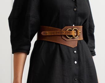 May include: A brown leather belt with a unique design. The belt features a wide, textured band with a brass buckle and two large brass rings. The belt is worn over a black dress.