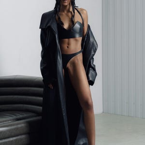 May include: A woman wearing a black leather coat, a black leather bra, and black underwear. She is standing in front of a gray wall and a black leather couch.