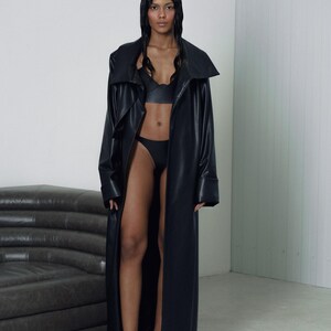 May include: A woman wearing a long black leather coat with a large collar. She is wearing a black bra and black underwear. She is standing in front of a gray couch.