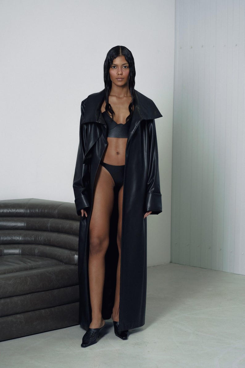 May include: A woman wearing a long black leather coat with a large collar. She is wearing a black bra and black underwear. She is standing in front of a gray couch.