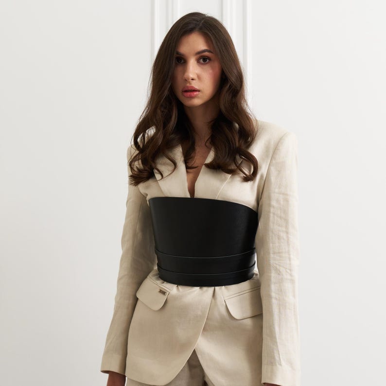 May include: A woman wearing a beige blazer and matching pants, paired with a wide, black leather corset. The corset cinches the waist, creating a defined silhouette. The outfit is set against a plain white background.