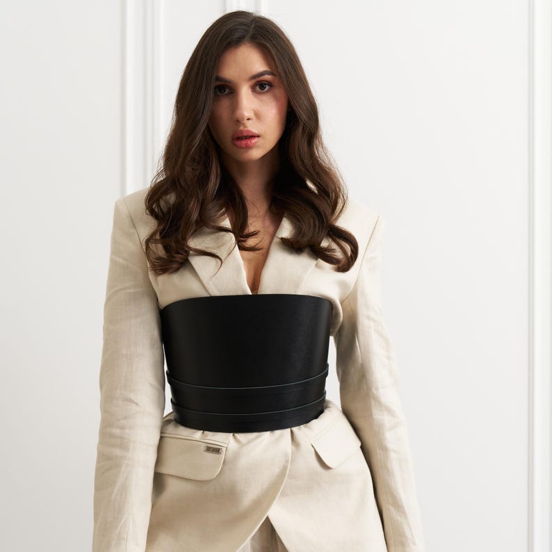 Black leather wide corset belt with structured panels and adjustable lace-up or buckle closure, designed to cinch the waist and enhance the silhouette.