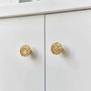 Peut inclure: Deux boutons de porte de placard de couleur or avec une texture martelée. Les boutons sont ronds et ont un bord légèrement surélevé.
