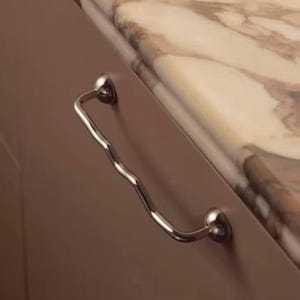 May include: A chrome cabinet pull with a distinctive wavy handle. The handle is fixed to the cabinet with two spherical mounts. The cabinet is a muted brown, and the countertop features a marble pattern with beige and brown tones.