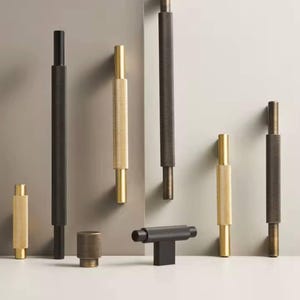 Könnte beinhalten: Eine Auswahl an Schrankgriffen und -knöpfen in verschiedenen Größen und Ausführungen. Die Hardware weist ein strukturiertes, zylindrisches Design in Schwarz, Gold und Bronze auf. Die Teile werden vor einem neutralen Hintergrund präsentiert.