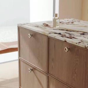 Op de afbeelding: Een houten kast met vier lades, voorzien van ronde metalen handgrepen. Het aanrecht is van marmer met bruine en witte aderen. Een parfumflesje en een tube crème staan op het aanrecht. De kast staat in een goed verlichte ruimte.
