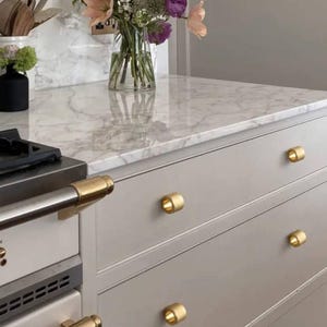 Może przedstawiać: Zbliżenie na zestaw szuflad z złotymi gałkami do szafek. Szuflady są jasnoszare i wykonane z drewna. Szuflady znajdują się na białym marmurowym blacie. Na blacie stoi wazon z kwiatami.