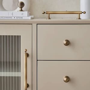 Pode incluir: Uma imagem de perto de um armário com ferragens de latão. O armário tem um acabamento bege claro e apresenta uma pega comprida, puxadores redondos e uma pega vertical. As ferragens têm um aspeto vintage.