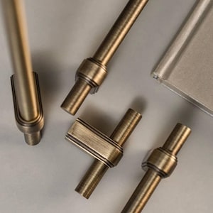 Può includere: Quattro stili diversi di ferramenta per armadi in oro spazzolato. La ferramenta include una maniglia a barra lunga, una maniglia a barra corta, una maniglia a forma di T e un piccolo pomo.