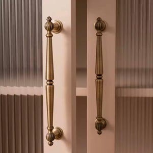 May include: Two antique gold cabinet door pulls with ornate details. The pulls are attached to a light pink cabinet door with a frosted glass panel.