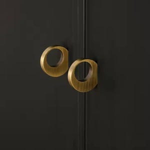 Puede incluir: Dos tiradores de armario de color latón con un diseño circular. Los tiradores están montados en una puerta de armario gris oscuro. Los tiradores tienen un acabado mate y una estética moderna. La puerta del armario está dividida por una línea vertical.