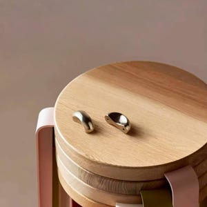 Op de afbeelding: Een houten kruk met een gelaagd ontwerp en een natuurlijke houten afwerking. De kruk heeft een ronde bovenkant en is versierd met twee zilveren metalen handvatten.