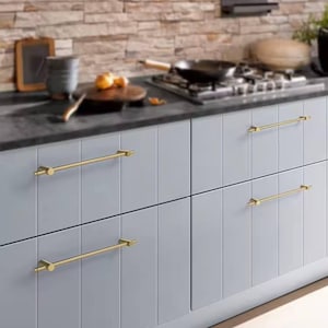Op de afbeelding: Lichtgrijze keukenkasten met goudkleurige stanggrepen. De kasten hebben verticale panelen en staan tegen een donker aanrechtblad en een bakstenen achterwand. De keukenomgeving omvat een kookplaat en kookgerei.