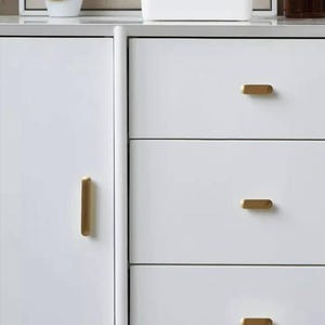 Pode incluir: Cômoda branca com três gavetas e puxadores retangulares dourados. A cômoda tem um design simples e moderno.