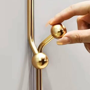 Puede incluir: Pomo de puerta de metal dorado con dos pomos redondos. El pomo está fijado a una pared blanca.