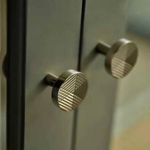 Könnte beinhalten: Zwei silberne Schrankknöpfe mit geometrischem Muster. Die Knöpfe sind rund und haben ein erhabenes Design.