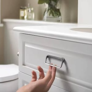 May include: A white bathroom cabinet with a silver rectangular drawer pull. The cabinet has a recessed panel design. A hand is pulling the drawer open. The background is blurred, showing a sink and decorative items.