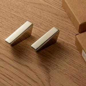 May include: Two polished, silver-toned cabinet pulls with a rectangular design. The pulls are angled and sit on a wooden surface. Two small, brown cardboard boxes are in the background.
