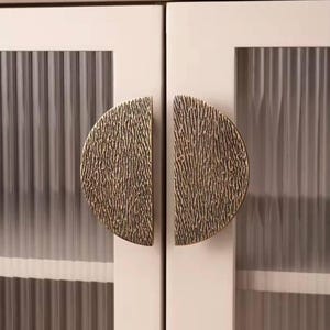 Puede incluir: Dos tiradores de puerta de armario en forma de semicírculo con un acabado en latón cepillado texturizado. Los tiradores están montados en una puerta de armario blanca con paneles de vidrio esmerilado.