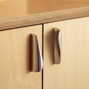 以下が含まれることがあります： 明るい木製のキャビネットドアに取り付けられた、2つのつや消しニッケルキャビネットプル。プルは湾曲した人間工学に基づいたデザインです。キャビネットドアは明るい色の木材で作られています。