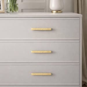 May include: A pale grey chest of drawers with three drawers, each with a textured gold handle. A white lamp with a gold base sits on top, along with a vase of white tulips. The chest of drawers is a piece of bedroom furniture.
